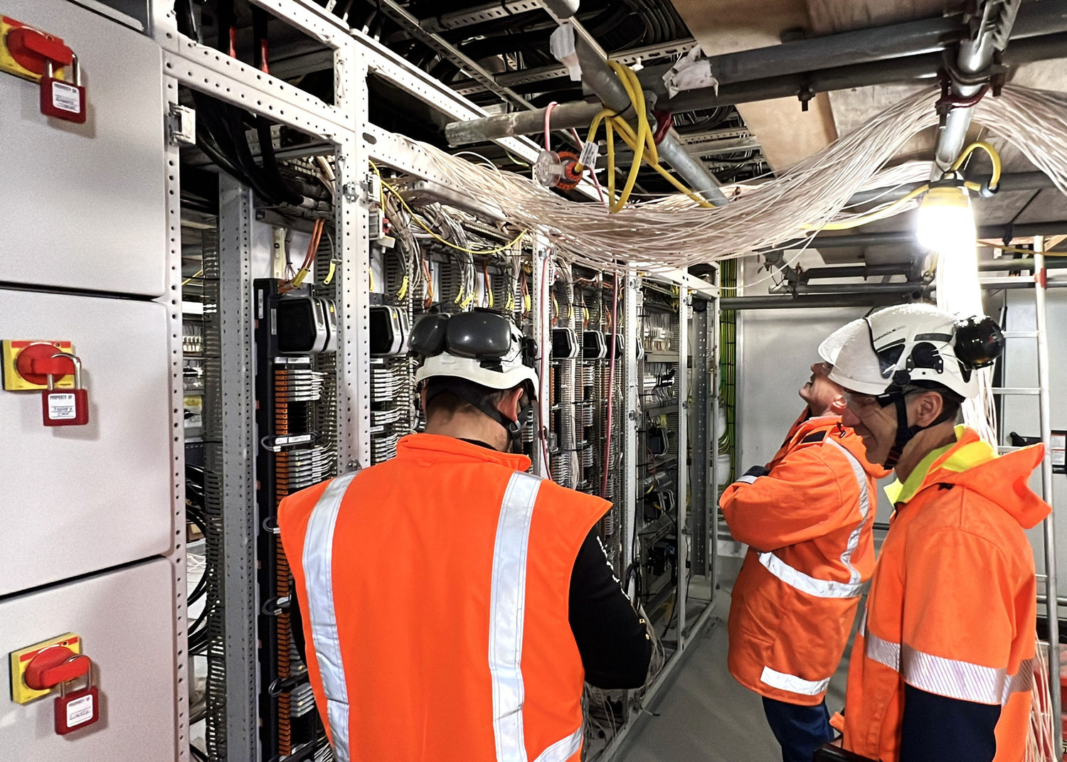 Three workers in orange vests and hard hats inspecting a server room.
