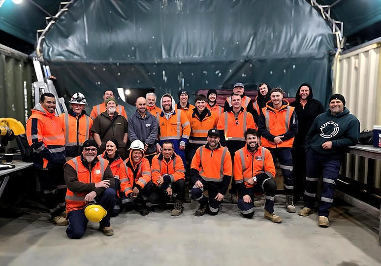 Group of people in orange vests and safety gear posing for a photo inside a large industrial or warehouse setting.